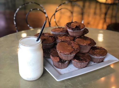 A plate of chocolate muffins next to a jar of milk with a straw, set on a table in a cozy café.