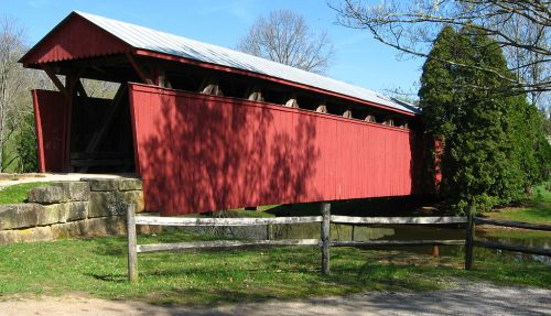 A red covered bridge stands over a small stream, surrounded by trees and a wooden fence on a sunny day.