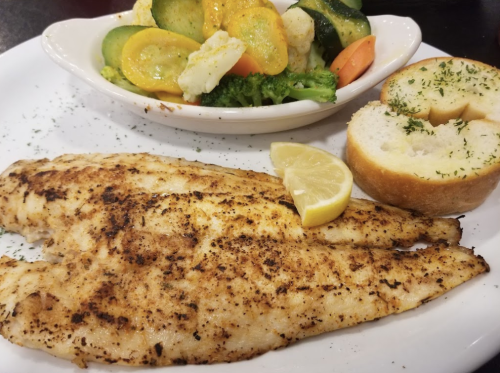 A plate featuring two fillets of seasoned fish, lemon wedges, a side of mixed vegetables, and a slice of garlic bread.
