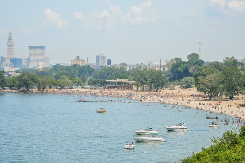 A busy beach with boats on the water, city skyline in the background, and people enjoying the sunny day.