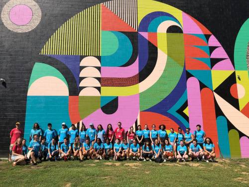 A diverse group of people in matching blue shirts poses in front of a colorful mural on a sunny day.