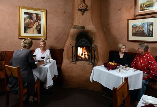 Two tables in a cozy restaurant with a fireplace, where four people enjoy dinner and wine in a warm atmosphere.