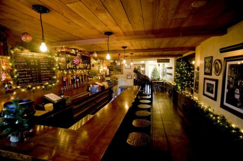 Cozy bar interior with wooden decor, festive lights, and a long counter lined with stools. Christmas decorations visible.