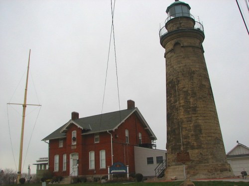 A tall lighthouse beside a brick building, set against a gray sky, with power lines in the foreground.