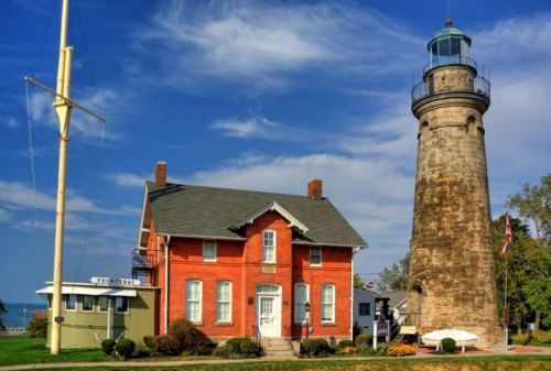 A historic red brick lighthouse and keeper's house beside a calm lake under a blue sky with scattered clouds.