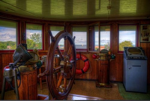 A vintage ship's wheel and navigational equipment inside a wooden cabin, with windows showing a scenic view outside.