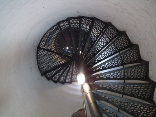 A spiral staircase viewed from above, featuring a black metal railing and a light source illuminating the steps.