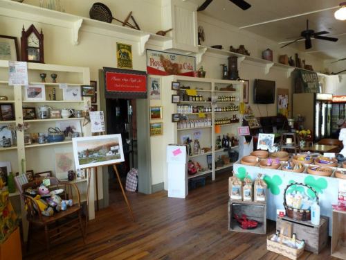 A cozy shop interior with shelves of goods, including jars, snacks, and vintage items, featuring wooden floors and ceiling fans.