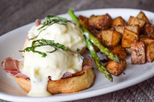 A plate of eggs Benedict with ham, hollandaise sauce, asparagus, and seasoned roasted potatoes.