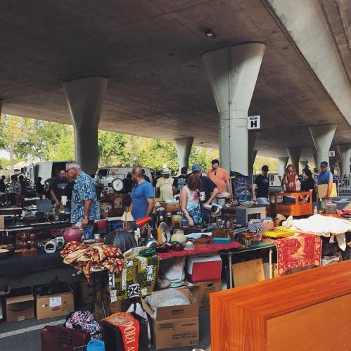 A bustling outdoor flea market with people browsing various antiques and vintage items under a concrete structure.