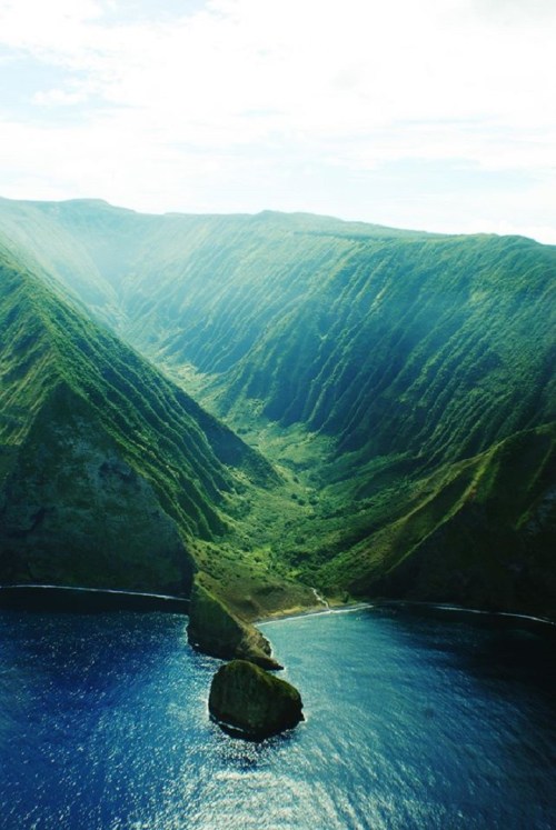 Aerial view of lush green mountains descending into a blue ocean, with a rocky outcrop in the foreground.