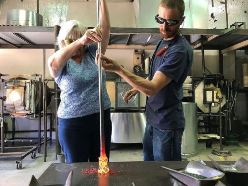 A woman and a man work together in a glassblowing studio, shaping molten glass with tools and protective eyewear.