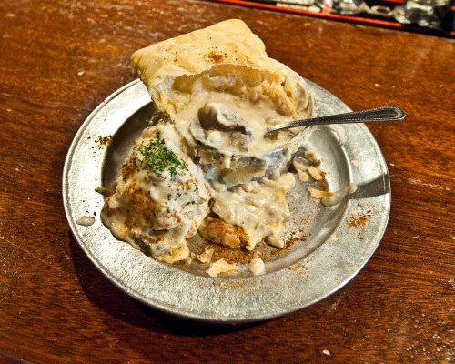 A silver plate with a flaky biscuit filled with creamy gravy and a side of mashed potatoes, garnished with parsley.