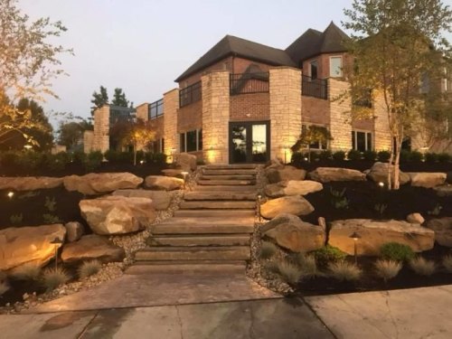 A modern house with stone accents, surrounded by landscaped rocks and plants, illuminated at dusk.