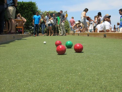 A vibrant outdoor scene with people socializing around a bocce ball court featuring red and green balls on the grass.