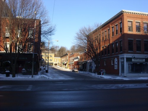 A quiet street scene in a small town, lined with brick buildings and snow, under a clear blue sky.