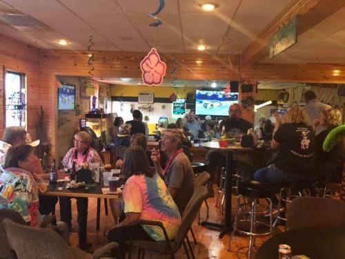 A lively bar scene with patrons chatting, colorful decor, and a TV showing a sports game in a cozy wooden setting.