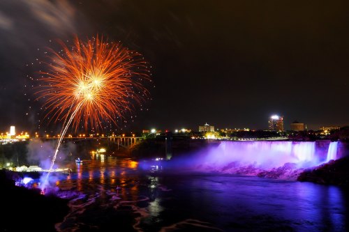 Fireworks illuminate the night sky over Niagara Falls, with colorful lights reflecting on the water below.