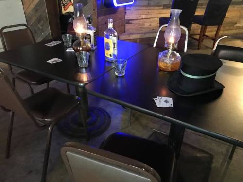 A dimly lit table with playing cards, glasses, a bottle, and a hat, set in a rustic bar environment.