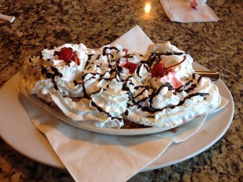 A dessert dish topped with whipped cream, chocolate drizzle, and cherries, served on a white plate with napkins.