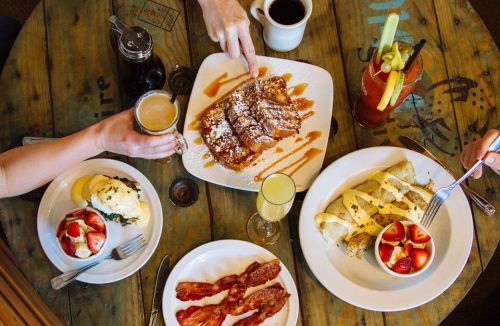 A table spread with various breakfast dishes: French toast, eggs Benedict, pancakes, bacon, and drinks.