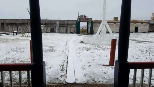 A snowy landscape viewed from a covered porch, with a path leading to a building in the background.