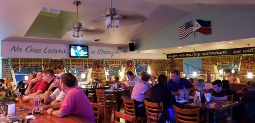 A cozy restaurant interior with patrons at the bar and tables, featuring flags and a welcoming sign on the wall.