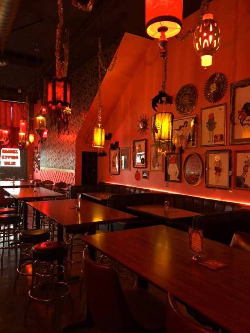 Cozy restaurant interior with red lighting, unique wall art, and wooden tables. Warm, inviting atmosphere.