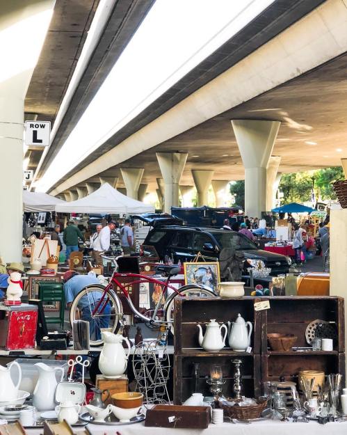 A bustling flea market under an overpass, featuring vintage items and shoppers exploring various stalls.