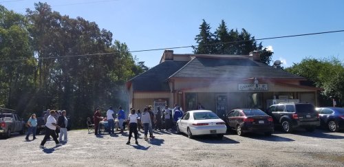 A busy market with people gathered outside, cars parked nearby, and smoke rising from a grill in the background.