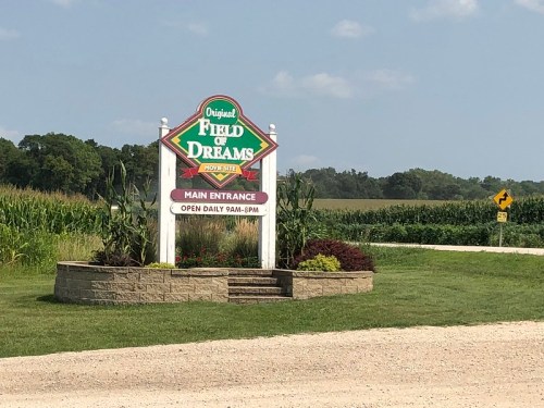 Sign for the Original Field of Dreams, featuring "Main Entrance" and hours, surrounded by green fields and blue sky.
