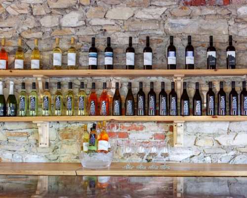 A wooden shelf displaying various bottles of wine against a stone wall, with glasses and ice in the foreground.