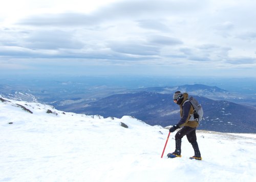 A person hiking on a snowy mountain, using an ice axe, with a vast landscape of hills and clouds in the background.