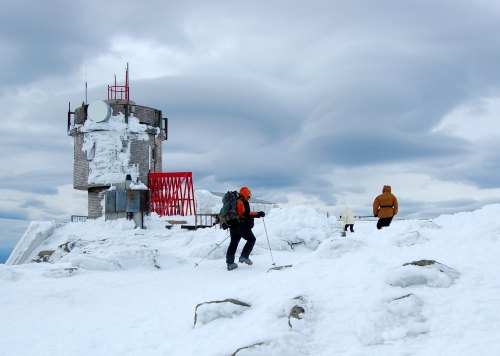 Two hikers navigate a snowy landscape near a weather station under a cloudy sky.