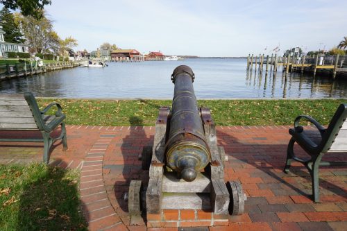 A historic cannon on a brick path overlooking a calm waterfront with boats and buildings in the background.