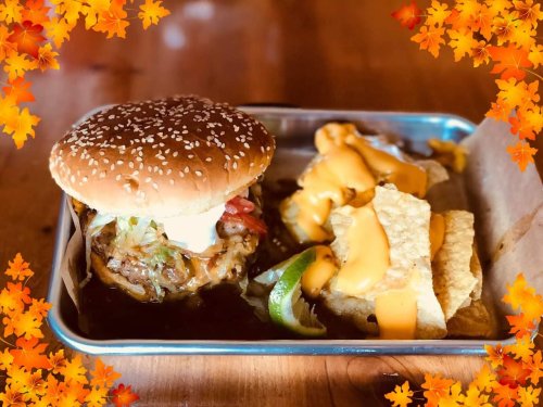A sesame seed burger with lettuce and tomato, served with nachos topped with cheese sauce, on a metal tray.