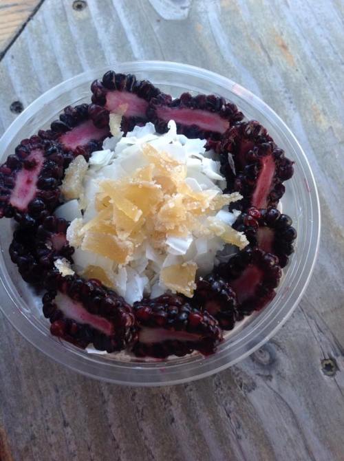 A clear cup filled with coconut flakes, topped with dark berries and pieces of candied ginger.