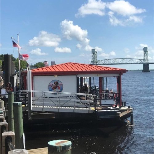 A riverside building with a red roof, docked by the water, with a bridge visible in the background and flags flying.
