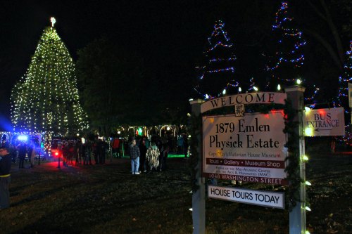 A festive night scene at the Emlen Physick Estate, featuring a large lit Christmas tree and visitors gathered outside.