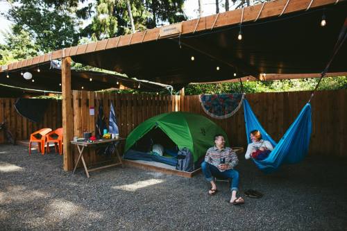 Two people relax in a campsite with a green tent, a hammock, and string lights under a wooden shelter.