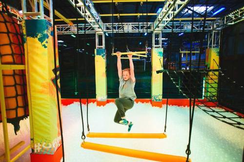 A child hangs from a bar in a colorful indoor obstacle course, surrounded by foam blocks and climbing structures.