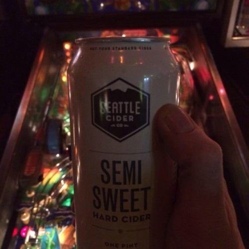 A hand holding a can of Seattle Cider Co. Semi Sweet Hard Cider in front of a colorful pinball machine.