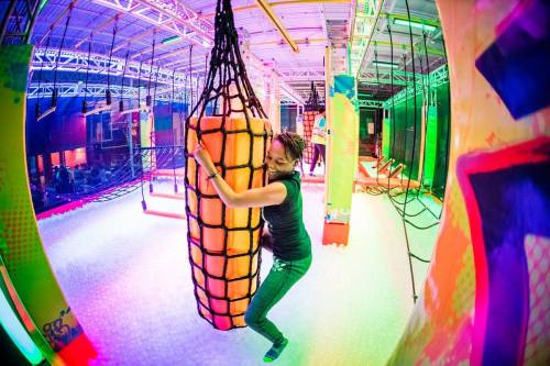 A person playfully hugs a large, colorful padded obstacle in a vibrant indoor adventure park.