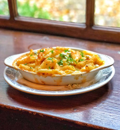 A creamy baked pasta dish topped with herbs, served in a white dish on a wooden table near a window.