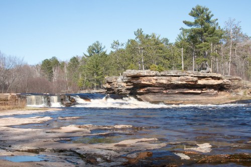 A serene river scene with a rocky outcrop, flowing water, and trees under a clear blue sky.