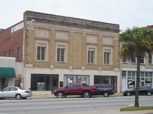 Historic brick building with decorative elements, for sale, flanked by smaller shops and parked cars on a street.