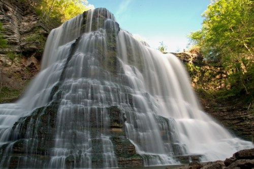 A majestic waterfall cascading down rocky cliffs, surrounded by lush greenery and a clear blue sky.