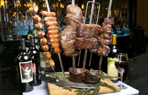 A display of various grilled meats on skewers, accompanied by red wine bottles and a glass of wine on a wooden board.