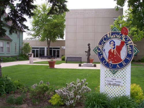 A park with a sign for "The Music Man Square," featuring greenery, benches, and a modern building in the background.