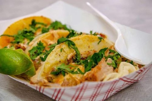 Three tacos filled with meat, topped with fresh cilantro, served with a lime wedge on a paper tray.